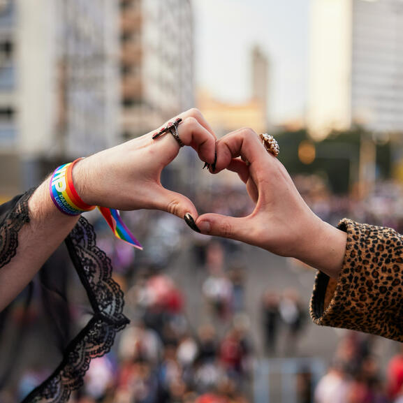 Photo de deux mains formant un coeur à une manifestation, un bracelet aux couleurs du drapeau LGBT visible sur le poignet de la main de gauche.
